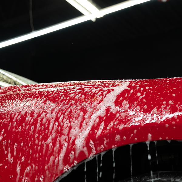 Red car with bubbly soap cascading down surface in a wash. Under bright lights, clean car detailing.